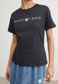 Person iført en sort GANT 1949 logo t-shirt og en blå denimnederdel med en trekantet lap på den nederste side.