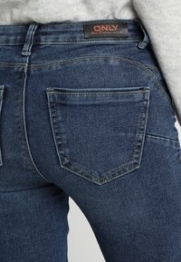 Tumma siniset denimfarkut, joissa on keskitason vyötärö, kaksi takataskua ja musta "ONLY"-logolla varustettu etiketti vyötäröllä. Kankaan pinta on teksturoitu.