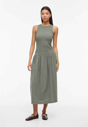 Naine seisab näoga edasi, seljas varrukateta oliivroheline midi-kleit, millel on hästi liibuv tekstuuriga ülakeha ja kergelt kootud seelik, kombineeritud tumedate madalate jalanõudega.