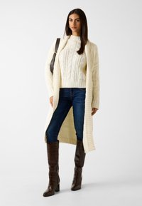 Romantische roomkleurige kabeltrui onder een lange roomkleurige cardigan, gecombineerd met donkerblauwe jeans en bruine kniehoge laarzen met hak en krokodillenstructuur.