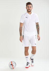 Polo shirt e pantaloni corti bianchi, con accenti neri. Calze bianche e scarpe con tacchetti con dettagli arancioni. Palla da calcio argentata nelle vicinanze.