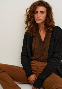 Dunkelgrauer, gestrickter Cardigan über einer braunen Bluse mit floralen Mustern und roten Akzenten, kombiniert mit braunen, figurbetonten Hosen. Texturierte Details im Stoff sichtbar.