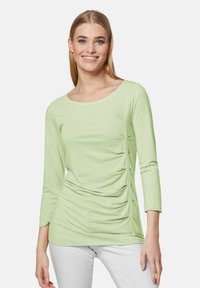 Hellgrünes langärmliges Oberteil mit Rundhalsausschnitt, seitlichen Raffungselementen und weicher Textur. Kombiniert mit weißen Hosen. Keine sichtbaren Muster.