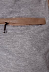 Šedá látka s hladkou texturou, doplněná hnědým koženým kapsou a zvýrazněná bronzovým jezdcem na zipu.