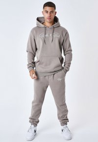 Grijze hoodie met witte logo, kangoeroezak en bijpassende grijze joggingbroek. Beide gemaakt van zachte stof, gedragen met witte sneakers.