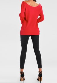 Femme portant un pull rouge à épaules dénudées, un jean skinny noir et des sandales à talons hauts noires, vue de dos sur un fond blanc.