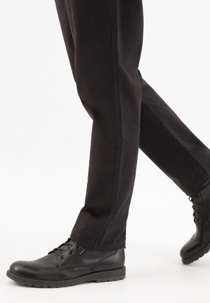 Zwarte leren veterschoenen gedragen met zwarte broek, getoond op een lopend persoon tegen een witte achtergrond.