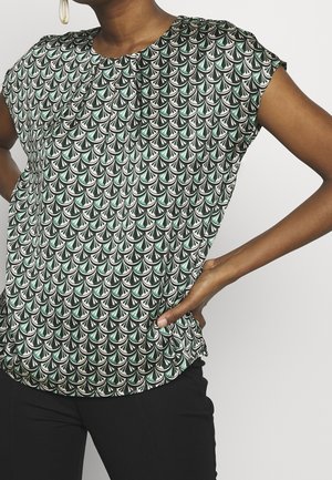 Femme portant un chemisier à manches courtes avec un motif écaille en vert, noir et blanc, associé à un pantalon noir et de grandes boucles d'oreilles créoles.