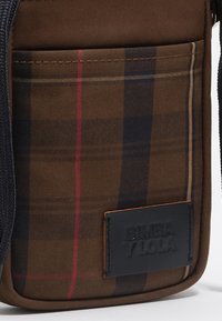 Bolso cruzado de cuadros marrones que presenta una etiqueta de cuero con texto en relieve. El diseño incluye detalles rojos, marinos y dorados en la tela.