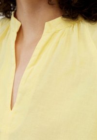 Żółta lniana bluzka z dekoltem w serek, z delikatną fakturą i lekko marszczonymi ramionami dla dodatkowego urozmaicenia.
