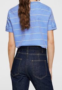 Chemise bleu à manches courtes avec des rayures blanches, dotée d'un col rond et d'un détail de bouton au dos, associée à un jean en denim foncé taille haute.