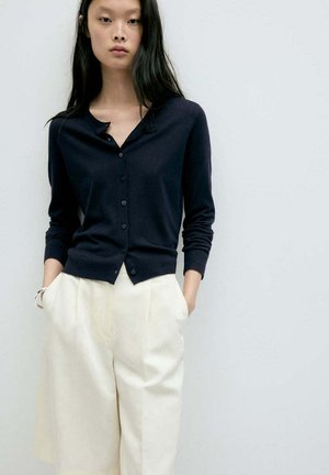 Femme aux longs cheveux foncés porte un cardigan bleu marine boutonné et un pantalon blanc ample, debout les mains dans les poches devant un fond uni.