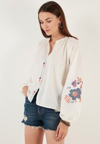 White linen blouse with a button-down front, featuring multicolored floral embroidery on sleeves and a slight puff at the cuffs.