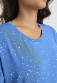 Niebieska bluzka z krótkim rękawem i dekoltem w szpic, z kropkowanym wzorem w kolorze różowym, białym i brzoskwiniowym na całej tkaninie. Miękki, lekki materiał.