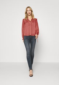 Blusa de color rojo oxido con estampado, con hombros fruncidos y cierre de botones, combinada con pantalones ajustados de mezclilla azul oscuro y tacones negros con tiras.