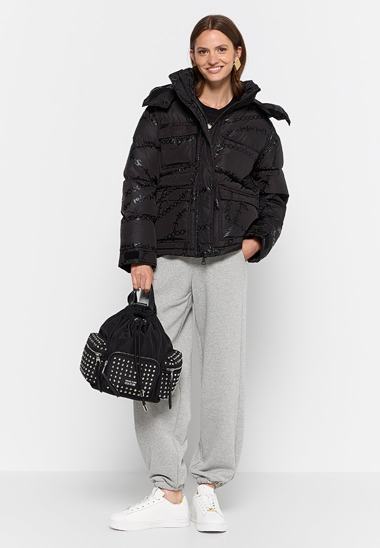 Chaqueta de plumas negra con material brillante, cierre de cremallera y bolsillos frontales. Combinada con pantalones de chándal grises y una mochila negra con tachuelas y cordón.