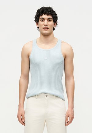 Νέος άνδρας με σγουρά μαλλιά που φοράει ανοιχτό μπλε ριμπ αμάνικη μπλούζα και εκρού παντελόνι, στέκεται μπροστά σε απλό λευκό φόντο.