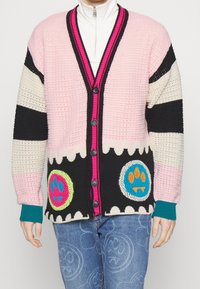 Kleurrijk gebreid vest met roze, zwarte en crèmekleurige strepen, voorzien van twee ronde applicaties en contrasterende geribbelde mouwen. Zwarte knopen sieren de voorkant.