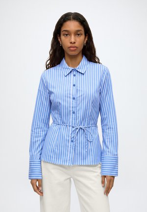Femme portant une chemise à rayures verticales bleues et blanches boutonnée avec un lien à la taille et un pantalon blanc, debout devant un fond uni.