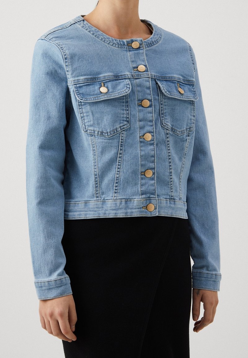Light blue denim jacket with round neckline, gold buttons down front, two buttoned chest pockets, worn over a black skirt.
