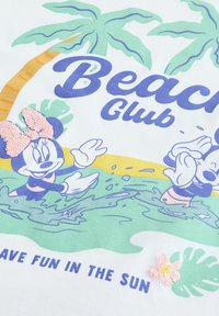Witte t-shirt met een kleurrijke "Beach Club" graphic, inclusief palmbomen, cartoonfiguren en paillettenaccenten in roze en groen.