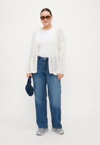 White crochet cardigan over a white top, paired with loose blue jeans. Accessories include a navy textured bag and grey sneakers.