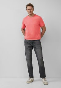 Korallenfarbene Baumwoll-T-Shirt mit kurzen Ärmeln, kombiniert mit abgetragenen dunkelgrauen Jeans und cremefarbenen Sneakers. Der Look zeichnet sich durch eine entspannte Passform und einen lässigen Stil aus.