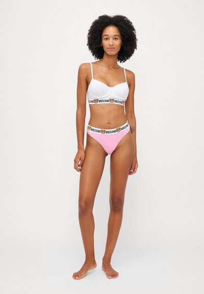 Bela bralette z tankimi naramnicami in blagovno označeno elastično stožčasto; rožnate bikini hlače z ujemajočim se blagovno označenim pasom; gladka tekstura tkanine.