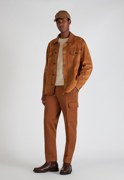 Braune Wildlederjacke mit Knopfverschluss, beiger Strickpullover, braune Cargo-Hosen mit Seitentaschen und dunkelbraune Stiefeletten.