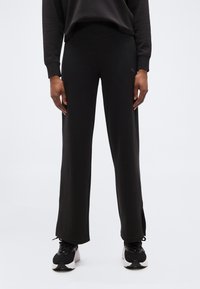 Άτομο που φοράει μαύρο παντελόνι με φαρδιά πόδια, μαύρο φούτερ και μαύρα και λευκά αθλητικά παπούτσια, στέκεται μπροστά σε απλό ανοιχτό φόντο.