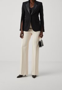 Czarna marynarka z satynowym kołnierzem, kremowe spodnie o szerokich nogawkach i czarne szpilki z ostrym noskiem. Trzyma małą czarną torebkę. Prosty i elegancki design.