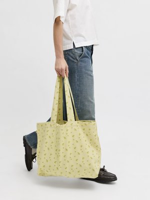 Osoba oblečená v modrých džínsoch a čiernych topánkach drží veľkú svetložltú tašku s drobnými zelenými kvetinovými vzormi.