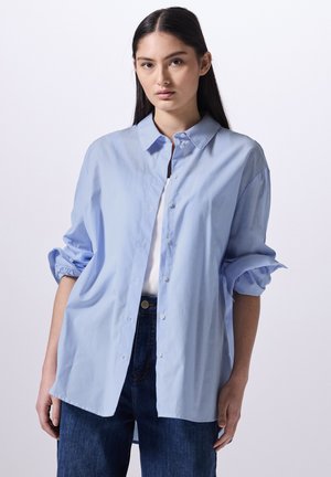 Camicia azzurro chiaro a righe con colletto, maniche arrotolate e una vestibilità ampia, abbinata a jeans in denim scuro.