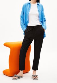 Camicia azzurra chiara con bottoni, t-shirt bianca sotto, pantaloni neri con cordino e ciabatte grigie; sedia scultorea arancione sullo sfondo.