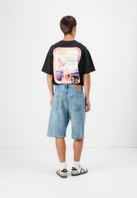 Svart t-skjorte med fargerik grafikktrykk på ryggen, kombinert med lyseblå denimshorts. Hvite sokker og svart-grå sneakers.