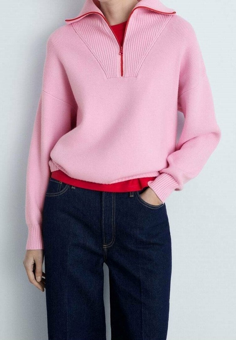 Personne portant un pull côtelé rose avec une demi-fermeture éclair sur une chemise rouge, associé à un jean bleu foncé, une main dans la poche.