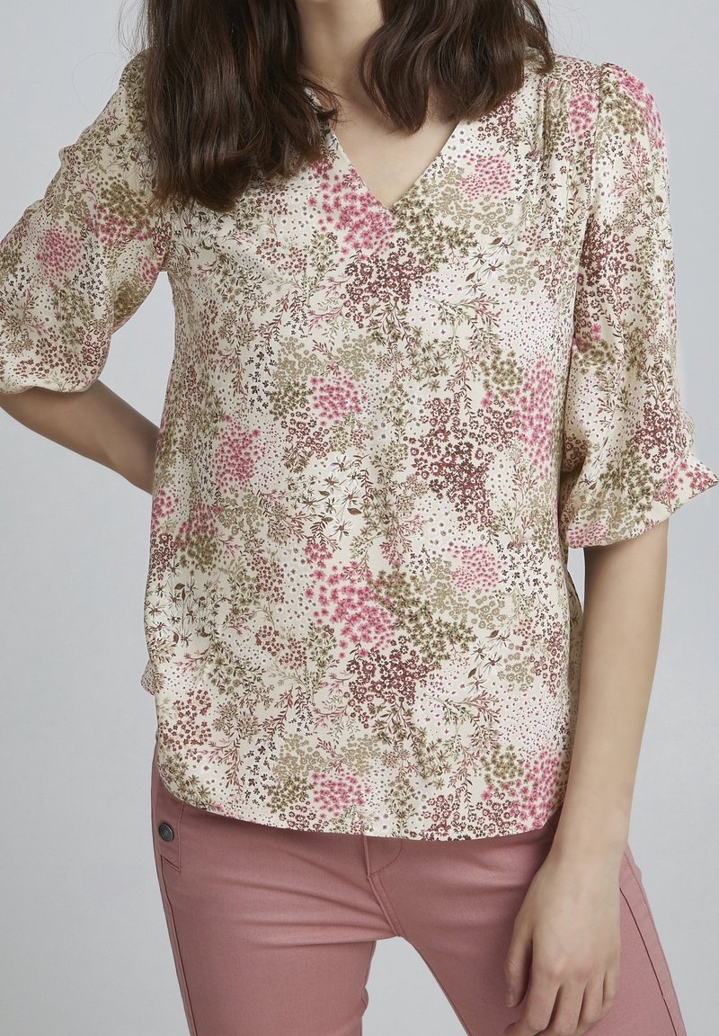 Blumenbluse in Creme mit rosa und grünen Mustern, mit V-Ausschnitt und kurzen Puffärmeln. Glatter Stoff mit leichter Textur.
