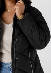 Zwarte gewatteerde pufferjas met een hoge kraag, ritssluitingen en drukknoopsluitingen, en zijzakken, gedragen over een gestreepte crèmekleurige top.