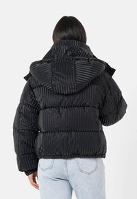 Person som bär en svart, glänsande, ribbad puffjacka med huva, sedd bakifrån, ihop med ljusblå jeans.