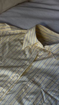 Chemise boutonnée jaune clair avec de fines rayures verticales bleues, posée à plat sur une surface grise texturée.