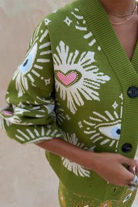 Cardigã verde tricotado com padrões brancos de olhos e explosões, detalhe de coração rosa com strass e botões pretos na frente.