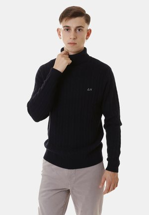 Maglione a collo alto blu navy a costine con una vestibilità aderente, caratterizzato da un piccolo logo sul petto e una trama in maglia. Abbinato a pantaloni chiari.