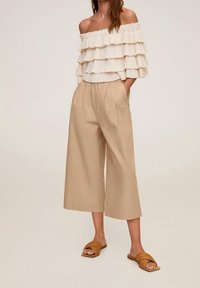 Vrouw in een off-shoulder crèmekleurige blouse met ruffles, beige wijd uitlopende cropped broek en bruine gevlochten instapsandalen, staande met handen in de zakken.
