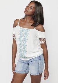 Top bianco off-shoulder con ricami floreali blu e dettagli in frange sulle maniche, abbinato a pantaloni corti in denim chiaro. Texture liscia.