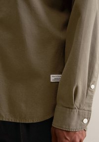 Koszula oliwkowozielona o gładkiej fakturze, z długimi rękawami i białymi guzikami. Z boku znajduje się metka z informacjami o marce.