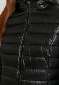 Černá prošívaná bunda s vysokým límcem a zipovým zapínáním. Má lesklou texturu a horizontální prošívaný vzor pro izolaci.