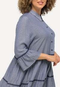 Pruhovaná button-up blůza v námořnické modré a bílé z bavlny. Obsahuje límeček, zvonové rukávy, dekorativní krajkové detaily na zakončení a rukávech.
