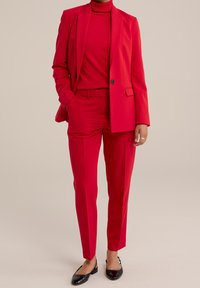 Costume rouge sur mesure avec un blazer et un pantalon slim assorti, associé à un col roulé rouge. Des ballerines noires complètent la tenue. Texture de tissu lisse.