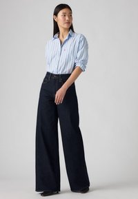 Lichtblauw gestreept overhemd met knopen, opgerolde mouwen; donkere wijdvallende jeans, donkerdenim; zwarte schoenen. Simpel ontwerp en duidelijke kleurcontrast.