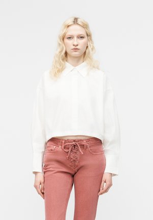 Femme blonde aux cheveux ondulés portant un haut court blanc à manches longues et un jean taille haute rose à lacets, posant devant un fond uni.